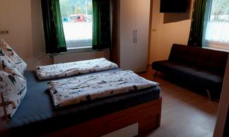 A cozy room with two beds, a sofa, and a large window. The walls are painted in neutral colors, and the windows are decorated with green curtains.