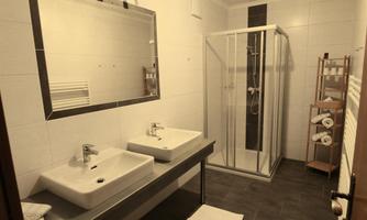 A modern bathroom with two sinks and a shower. The space is bright and clean, with a towel rack in the corner.