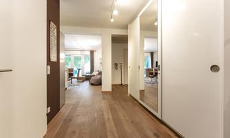 A modern hallway with wooden flooring and large mirrors. In the background, there are living furnishings and windows with a view of the outdoors.