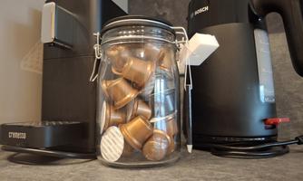 A modern kitchen corner with a coffee capsule machine and a kettle. A glass container with coffee capsules is in the foreground.
