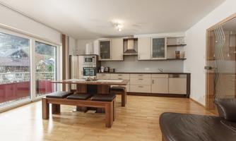 A modern kitchen with wooden flooring and large windows. The dining table is made of wood and accommodates several people.