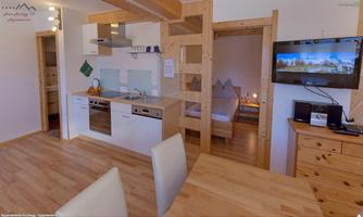 A modernly furnished apartment with an open kitchen and a dining area. In the background, a bedroom and a television are visible.