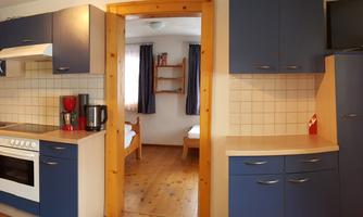 A modern kitchen with blue cabinets and wooden details. In the background, you can see a bedroom with windows and bright light.