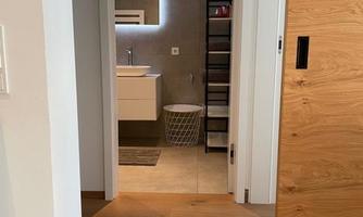 A bright hallway with wooden flooring that leads to a modern bathroom. The bathroom is minimally furnished, with a sink and a large wall mirror.