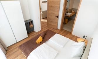 A cozy bedroom with a large bed and wooden floor. The room is bright and modern, with an open door to an adjacent bathroom.
