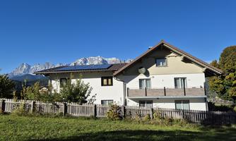 A beautiful house surrounded by green spaces and trees. In the background, the mountains are visible under a clear blue sky.