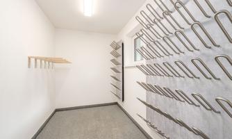 A bright room with white walls and a gray floor. Several metal hooks and a wooden shelf are installed on the wall.