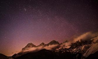 An impressive mountain landscape under a clear night sky full of stars. Gentle wisps of fog wrap around the peaks of the mountains.