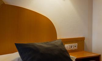 A cozy bedroom with a bed and a nightstand. A lamp hangs on the wall, providing warm light.