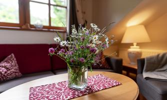 A beautiful bouquet of flowers in a glass vase is on a table. In the background, cozy seating furniture and a lamp can be seen.