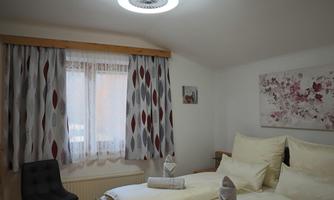 A cozy bedroom with two beds and bright pillows. Large windows with curtains and decorative elements on the walls create an inviting atmosphere.