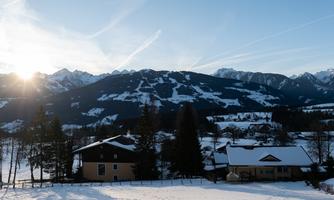 A winter landscape with snow-covered hills and mountains in the background. Two charming houses stand in the quiet, snowy surroundings.