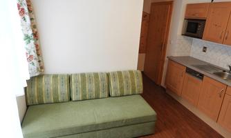 A cozy living room with a green sofa and a small kitchen in the background. The walls are bright and the windows have colorful curtains.