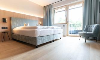 A modern bedroom with a large bed and a cozy armchair. Plenty of light comes through the windows and the floor is made of light wood.