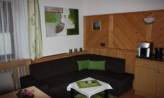 A cozy living room with a dark gray couch and green cushions. Modern artworks hang on the wall, while a small table and a coffee maker complete the ambiance.