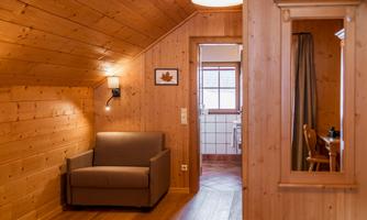 A cozy room with wooden paneling and a comfortable armchair. In the background, a bathroom and a mirror are visible.