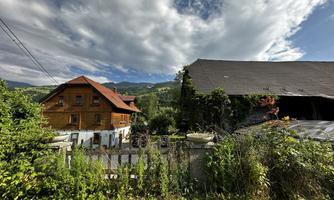 A rustic wooden farmhouse with a large roof. In the background, green hills and a cloudy sky can be seen.