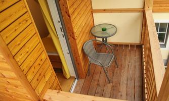 A small wooden balcony with a table and a chair. In the background, a bedroom is visible.