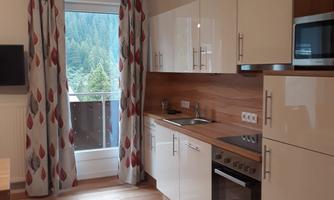 A modern kitchen with white cabinets and wooden details. The room has large windows that offer a view of nature.