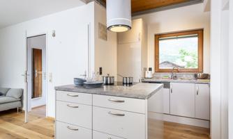 A modern kitchen with a central cooking island and wooden flooring. Large windows provide plenty of daylight and a view outside.