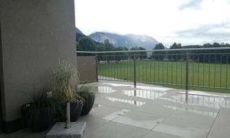 A modern terrace with gray tiles and plants. In the background, green fields and mountains are visible.