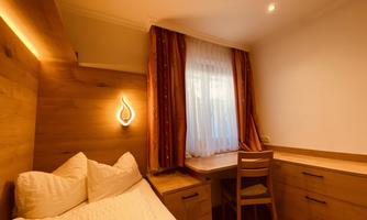 A cozy room with a single bed and a desk. Large windows let in plenty of light, while wooden shelves create a warm atmosphere.