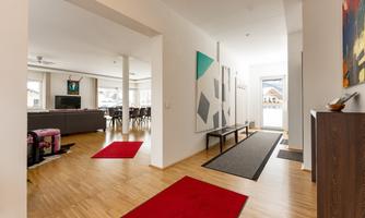 A modern hallway with light wood flooring and red carpets. In the background, a living area and large windows are visible.