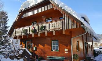 A cozy wooden house in winter with snow on the roof. The balcony is decorated with plants and the surroundings are covered in snow.