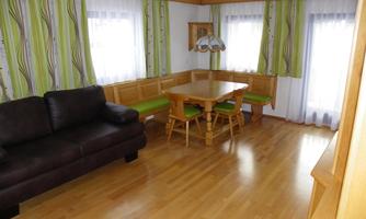 A cozy room with a dark sofa and a wooden dining area. Large windows let in plenty of light and the walls are adorned with green curtains.
