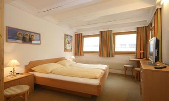 A simple hotel room with a double bed and wooden furniture. The windows are adorned with colorful curtains and there is a television as well as a desk.