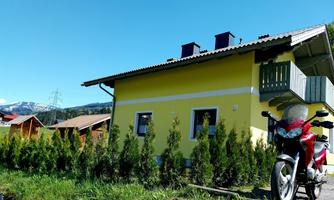 A yellow house with a balcony in a quiet environment. In the foreground, there is a motorcycle, and in the background, mountains can be seen.