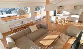 A modern living room with a comfortable sofa and a wooden table.  
Large windows provide plenty of daylight and a view of the terrace.