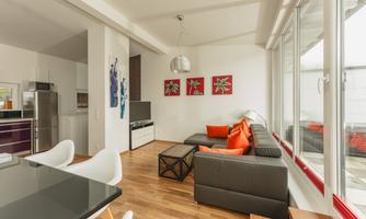 A modern living room with a gray sofa and orange cushions. The room has large windows and a stylish dining table.