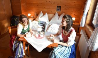 Two women in traditional costumes are sitting at a table in a cozy wooden room. They are enjoying a cup of coffee and smiling at each other.