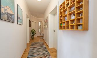 A bright hallway with wooden floors and a colorful carpet. Pictures hang on the wall, and a shelf with decorative figures is visible.