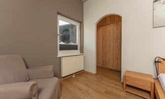 A simple room with a cozy armchair and a wooden wardrobe. The window lets in a lot of daylight and has a view outside.