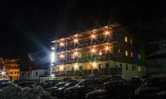A multi-story building illuminated at night, surrounded by snow and many parked cars. The facade has colorful windows and radiates an inviting atmosphere.