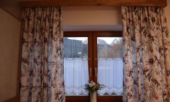 A window with floral curtains and a small vase with flowers on the windowsill. In the background, trees and a building are visible.