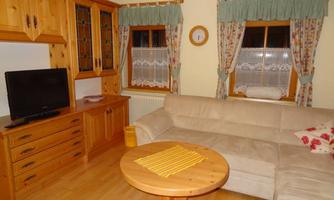 A cozy living room with a beige sofa and a wood-colored coffee table. The walls are painted in soft colors and large windows with curtains provide daylight.