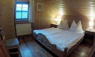 A cozy wooden bedroom with a double bed and two nightstands. The room has large windows and a warm, inviting atmosphere.