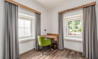 A bright room with two windows and gray curtains. In the middle is a wooden table with a green chair.
