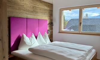 A modern bedroom with a double bed and a purple wall covering. Large windows provide plenty of daylight and a cozy atmosphere.