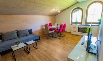 A modern living room with a cozy couch and a dining table with pink chairs. Large windows ensure plenty of light and a pleasant atmosphere.