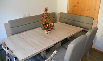 A cozy dining area with a wooden table and beige benches. On the table sits a small Christmas tree with lights and decorations.