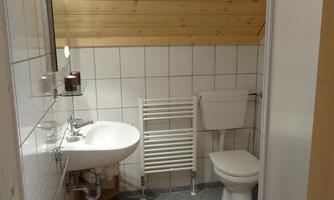 A small bathroom with bright tiles and a wooden ceiling. There is a sink, a toilet, and a shower cabin, complemented by an orange carpet.