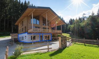A modern wooden house surrounded by green meadows and trees. The sun shines clearly over the building.