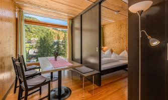 A modern room with wooden walls and a large window that offers a view of the mountains. There is a table with chairs and a cozy bed in the background.