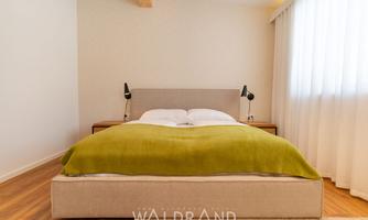 A cozy bedroom with a modern bed and a green blanket. The room has large windows that let in a lot of light.