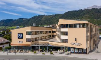 A modern hotel building with a bar and terrace area. The surroundings are surrounded by green mountains.