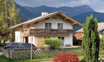 A modern house with a balcony, surrounded by trees and a beautiful garden. In the background, mountains are visible, complementing the landscape.